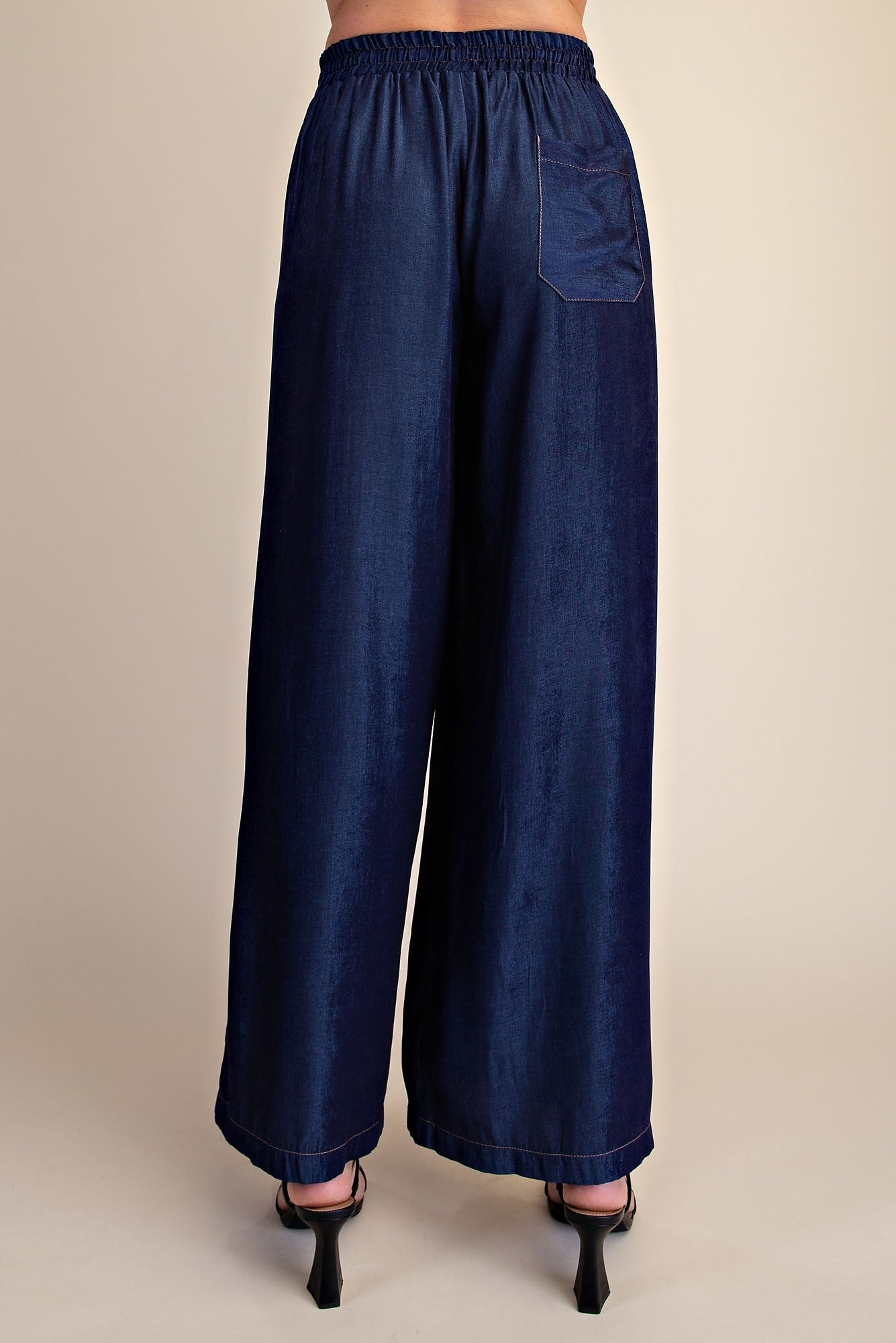 A person shown from the back wearing dark blue wide-leg pants with an elastic waistband and a side pocket, paired with black high-heeled sandals against a plain beige studio background.