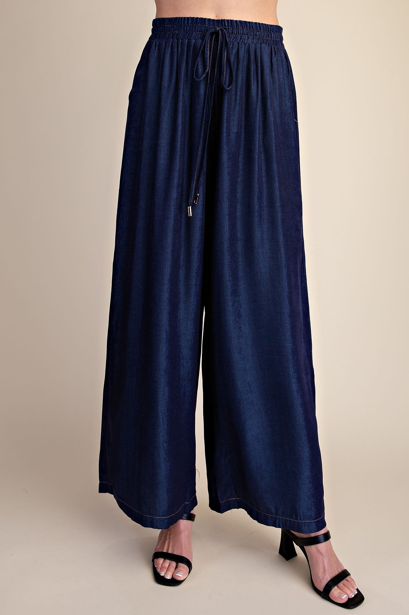 A person wearing dark navy wide-leg drawstring pants with black heeled sandals, shown from the waist down against a plain beige studio background.